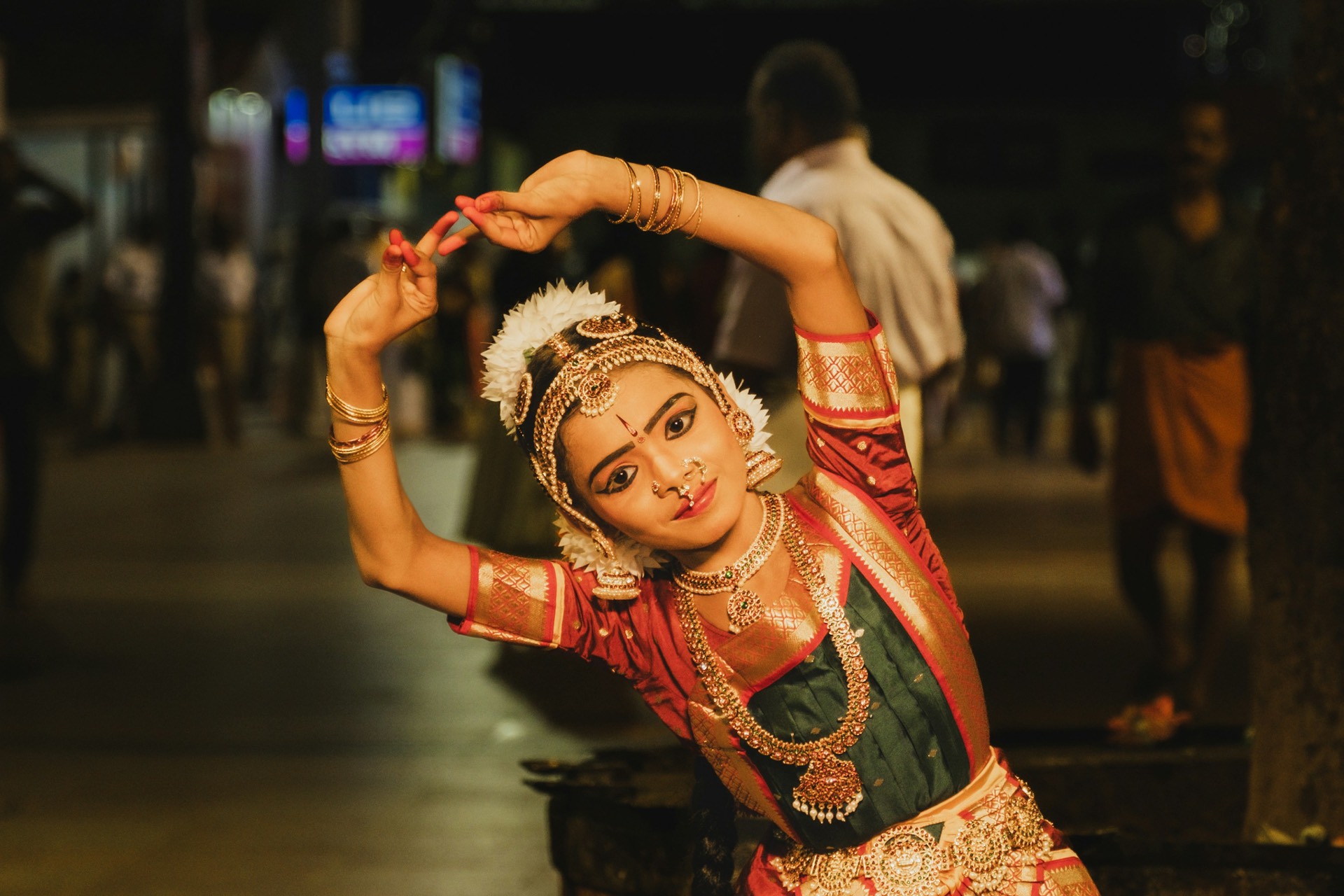 A Bharatanatyam dancer mid-pose in traditional costume with gold jewelry and stage makeup, hands raised in mudra